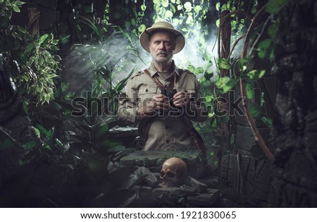 Confident explorer walking in the jungle, he is standing next to ancient ruins and holding binoculars