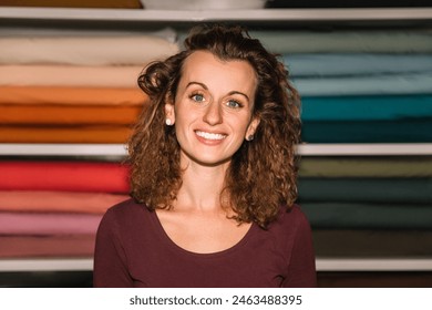 A confident fashion designer with curly hair poses in front of her well-organized colorful fabric rolls, exuding a professional and cheerful demeanor in her studio - Powered by Shutterstock