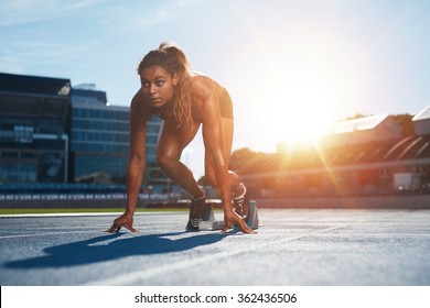 Confident Female Athlete In Starting Position Ready For Running. Young Woman About To Start A Sprint Looking Away With Bright Sunlight From Behind.