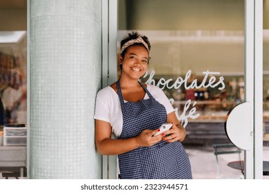 Confident female cafe owner, smiling while using her smartphone to manage and run her small restaurant. Female small business owner achieving success in her entrepreneurship. - Powered by Shutterstock