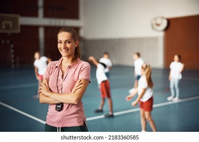 Confident Female Coach During Physical Education Class At School Gym Looking At Camera. Group Of Kids Are Exercising In The Background. 