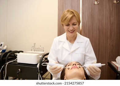 Confident Female Doctor Cosmetologist, Aesthetician Doing Professional Skin Care Procedures To The Face Of A Woman Receiving Stimulating Electrical Facial Microcurrent Therapy In Wellness Spa