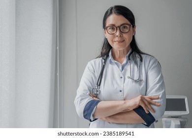 Confident female doctor standing at office and smiling at camera. Successful female doctor nurse medical worker caregiver gynecologist pediatrician with arms crossed ready to help assist