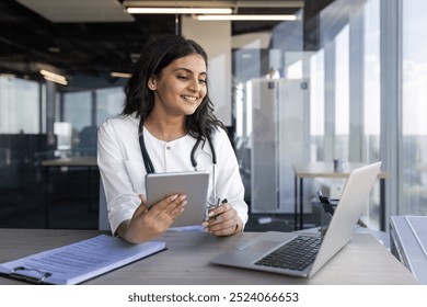 Confident female doctor with stethoscope engaged in online consultation using digital tablet and laptop. Healthcare professional smiling, offering virtual medical advice in modern office environment - Powered by Shutterstock