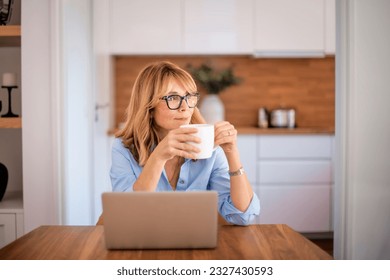 Confident female drinking tea while sitting at table at the kitchen. Mid aged woman using laptop for work. Home office. 