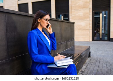 Confident Female Employer Talking Via Mobile Pone And Writing Notary In Textbook While Sitting Outside Enterprise During Break At Job 