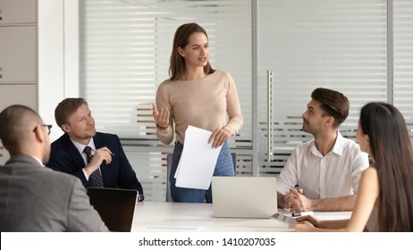 Confident female manager presenting financial report speak at team meeting talk to employees group, business woman company executive leader hold papers explain new business plan at corporate briefing