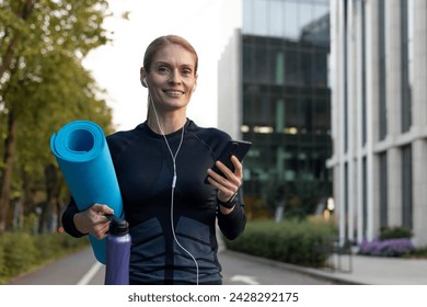 Confident female professional enjoying workout break, holding yoga mat and water bottle, using phone outdoors. - Powered by Shutterstock