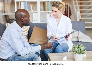 Confident Female Psychotherapist In White Coat Sitting On Sofa And Showing Records To Patient While Explaining Reason Of His Abnormal Behavior