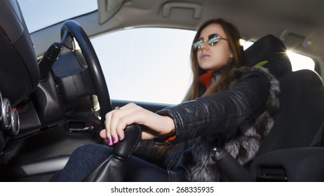 Confident Female Racer Drives Car. Young Woman Drives Car, Focus On Her Hand Switching Transmission Grip Biker Dress Black Leather Jacket, Sunglasses, Bright Sunbeams On Right Corner