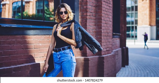 Confident female student with folder walking in campus in break of lessons, half length portrait of young caucasian hipster girl with jacket looking at camera during free time on urban setting
