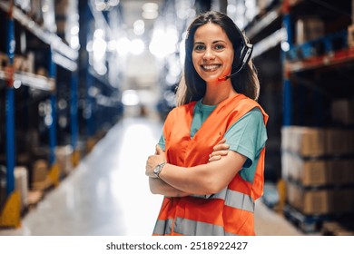 A confident female warehouse worker stands with arms crossed, wearing a headset in a brightly lit industrial space, symbolizing confidence and readiness in logistics work. - Powered by Shutterstock