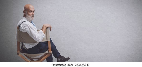 Confident Film Director Sitting On The Director's Chair And Looking At Camera: Video Production, Filmmaking And Film Industry Concept