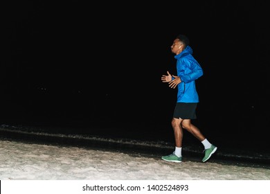 Confident Fit Young African Sports Man Wearing Windbreaker Exercising Outdoors At Night Time, Running At The Beach