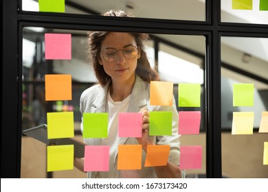 Confident focused businesswoman wearing glasses writing ideas or tasks on sticky papers on glass wall, female team leader, executive manager holding computer tablet, planning project, organize work