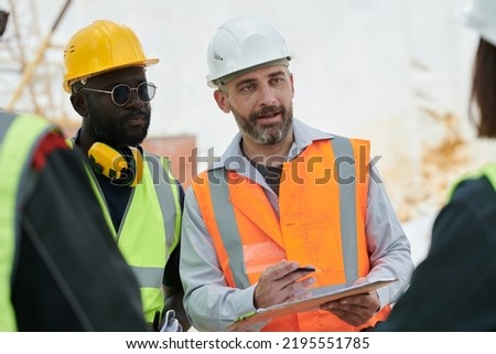 Confident foreman with document and pen explaining something to subordinates or giving instructions while looking at one of them