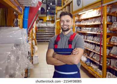 Confident friendly man working in warehouse in hypermarket of building materials. Smiling young male worker in coveralls standing with folded arms in one of aisles between shelves with goods.