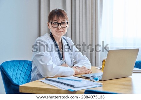 Confident friendly mature woman doctor sitting at table with laptop computer