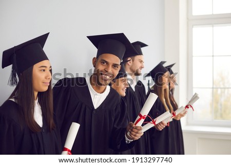 Confident in future. Smiling African-American exchange program student in black graduation cap and gown holding university diploma and looking at camera, standing together with international graduates