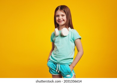 Confident Girl In Stylish Summer Outfit, Wearing White Headphones On Neck, Keeping Hands In Pockets, Isolated Over Yellow Brick Wall