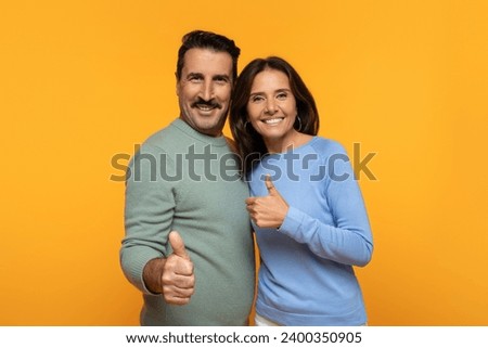 Confident glad old european wife and husband hugs, show thumbs up gesture with hands, approval and positivity on orange background. Approval and recommendation, ad and offer