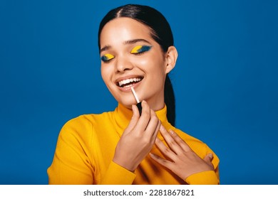 Confident and glamorous young woman applying lip gloss on her lips during her beauty routine. Woman with vibrant eyeshadow smiles as she finishes up her look with a glossy lip.  - Powered by Shutterstock