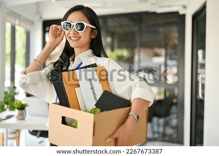 Confident and gorgeous millennial Asian female office worker, wearing sunglasses and holding a cardboard box with her belongings, feels happy to quit her job.