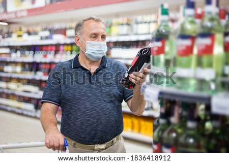 Confident grey-haired elderly man in protective mask choosing white wine in modern wineshop