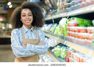 Confident grocery store employee with crossed arms smiling in front of a well-stocked vegetable aisle. Symbolizes customer service, retail work, and a positive working environment in a supermarket. - Powered by Shutterstock