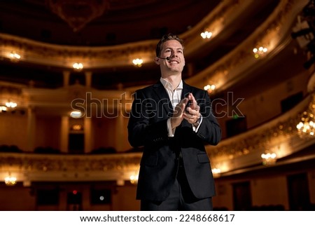 Confident guy of american appearance in formal classic suit clapping hands after performance, giving speech, on stage. Presenter Speaking to Audience People. Unidentifiable Audience and Presenter.