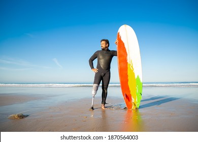 Confident handicapped man standing on sea beach with board. Attractive brunette man with artificial leg wearing black wetsuit and looking at ocean. Physical disability and extreme sport concept - Powered by Shutterstock