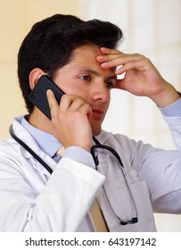 Confident Handsome Doctor With An Stethoscope Around His Neck, Using A Celphone With One Hand And With His Other Hand He Is Touching His Head, In Office Background