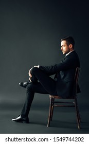 Confident Handsome Man In Black Suit Sitting On The Chair On Dark Backround. Profile View Full Lenght Male Portrait