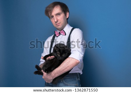 confident handsome man in shirt, suspender and bow tie holding black cat and looking at camera on blue background with copy space