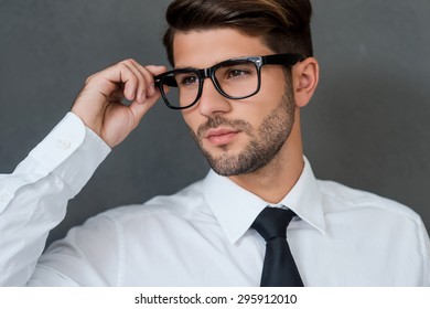 Confident and handsome. Confident young businessman adjusting his eyeglasses and looking away while standing against grey background - Powered by Shutterstock