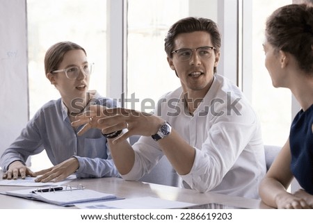Confident handsome young professional man speaking to female colleagues on brainstorming meeting, telling business project idea for discussion. Boss, team leader, mentor explaining task to employees