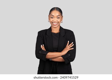 Confident happy African American businesswoman in a sleek black suit stands with arms crossed, smiling assuredly against a neutral gray background, studio. Work, business - Powered by Shutterstock