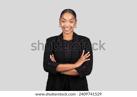 Confident happy African American businesswoman in a sleek black suit stands with arms crossed, smiling assuredly against a neutral gray background, studio. Work, business
