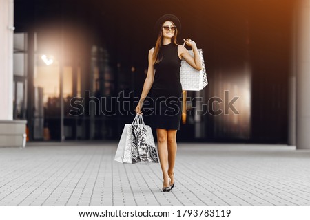 confident, happy, attractive young woman in a black dress and hat, wearing dark glasses, holding shopping bags in front of a shopping center. Black Friday, shopping