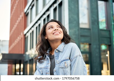 Confident happy beautiful young hipster African American woman wearing denim jacket looking up standing on city street outdoors dreaming, thinking or good future on urban buildings background.