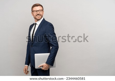 Confident happy businessman in eyeglasses, suit holds laptop looking on copy space through shoulder on grey background. Business, office work, leader and boss concept. Advertisement banner, poster. [[stock_photo]] © 