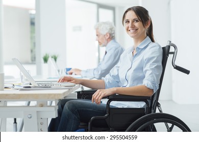 Confident Happy Businesswoman In Wheelchair Working At Office Desk And Using A Laptop, She Is Smiling At Camera, Disability Overcoming Concept