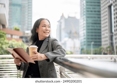 A confident, happy mature Asian businesswoman in a formal business suit stands on a skywalk with a book and a takeaway coffee cup in her hand, looking away at the skyscraper with a dreamy face. - Powered by Shutterstock