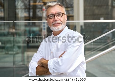 Confident happy mature older business man leader, smiling middle aged senior old professional businessman wearing white shirt glasses crossed arms looking at camera standing outside, portrait.