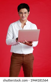 Confident Happy Young Asian Business Expert Handsome Man With Glasses In White Shirt Holding Computer Laptop And Typing Smiling Looking While Standing Against Red Wall  Background.  South East Asia