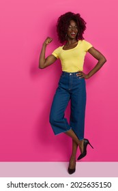 Confident And Happy Young Black Woman In Yellow Top, Jeans And High Heels Is Standing On One Leg, Flexing Muscles And Laughing. Full Length Studio Shot On Pink Background.