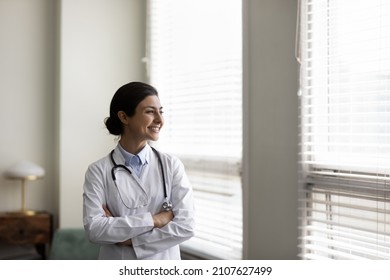 Confident happy young Indian woman GP doctor therapist physician cardiologist in white medical coat standing with folded arms in clinic office, looking in distance thinking of illness treatment.