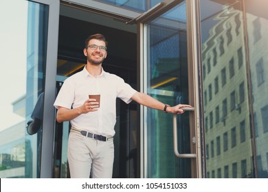 Confident Happy Young Man Opening The Door And Drinking Coffee While Walking Out From The Modern Office Building, Copy Space. Business, People And Success Concept