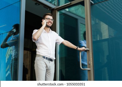 Confident Happy Young Man Opening The Door And Talking On Cell Phone While Walking Out From The Modern Office Building, Copy Space. Business, Technology And Communication Concept