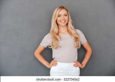 Confident in her abilities. Smiling young blond hair woman holding hands on hips and looking at camera while standing against grey background - Powered by Shutterstock